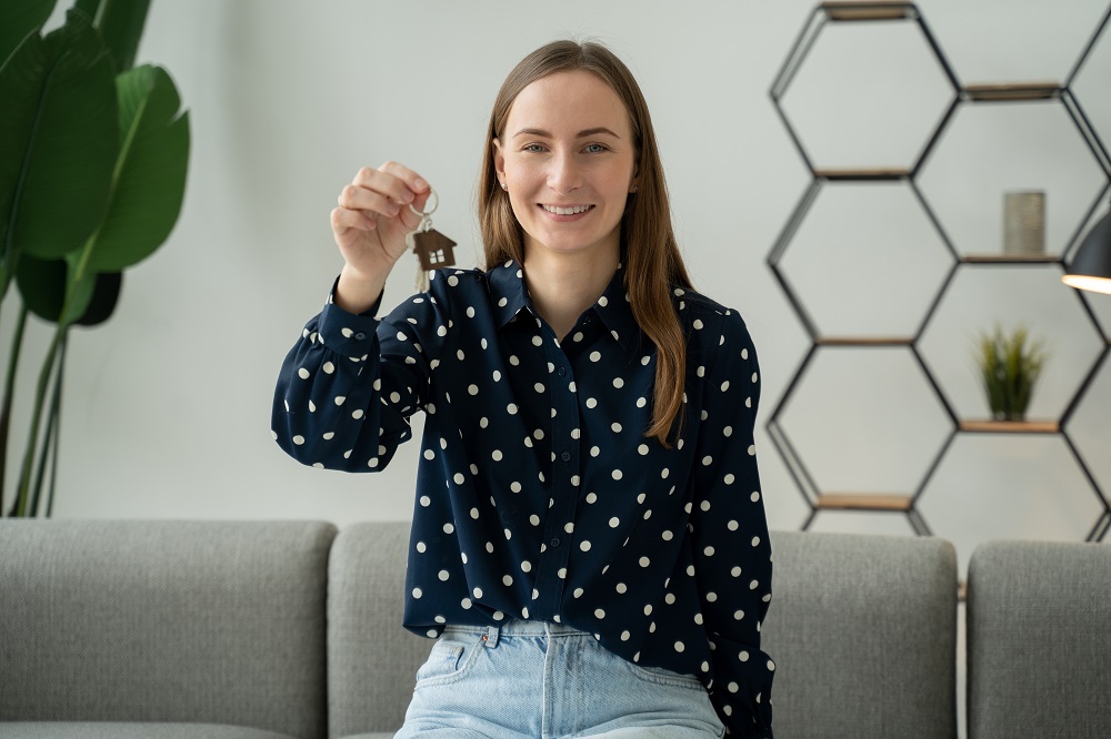 Uśmiechnięta, młoda kobieta wyciąga przed siebie rękę, w której trzyma klucz z brelokiem w kształcie domu